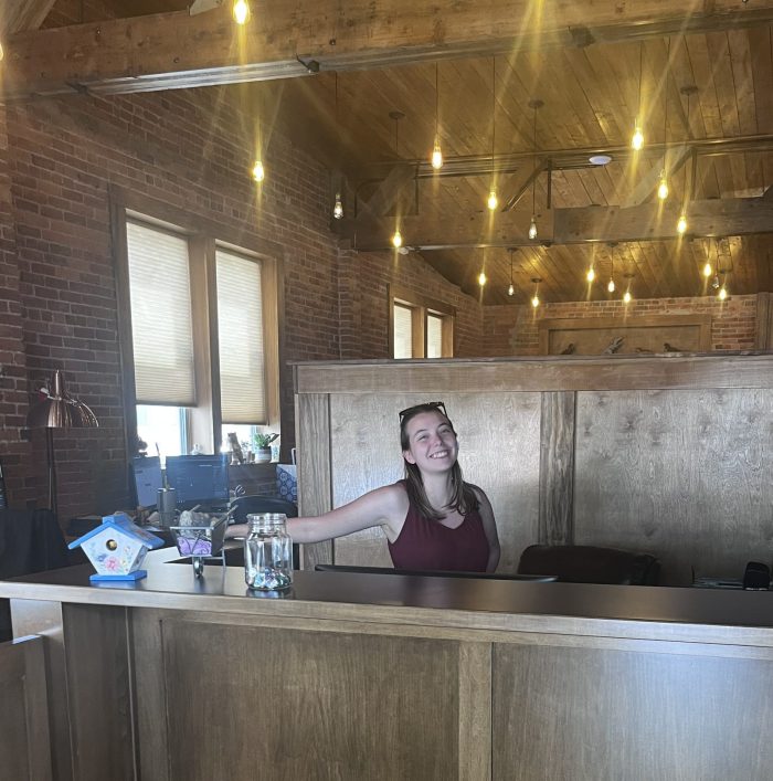 A smiling woman stands behind a wooden desk in an office with a high ceiling and brick walls.