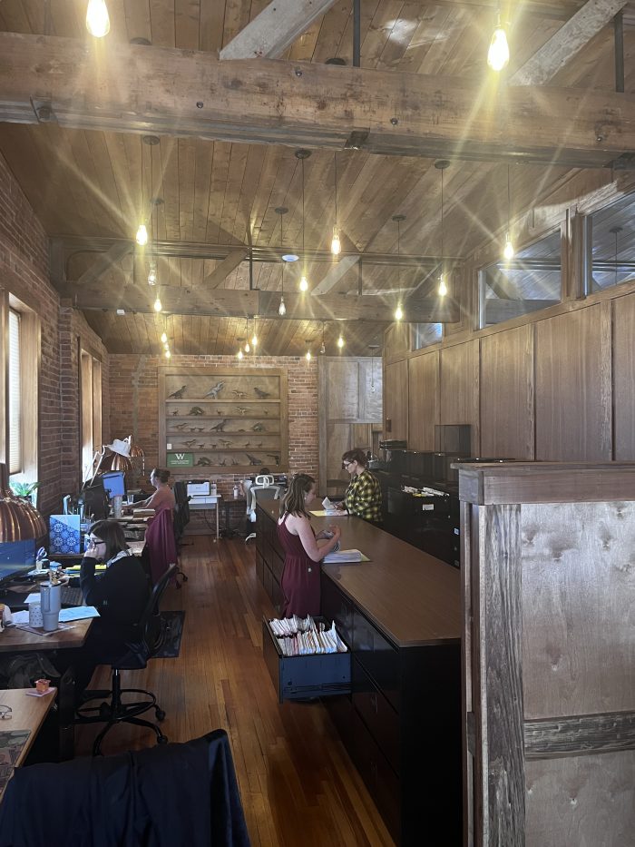 Employees working at computers and sorting papers in an office with long wooden desks and paneling.